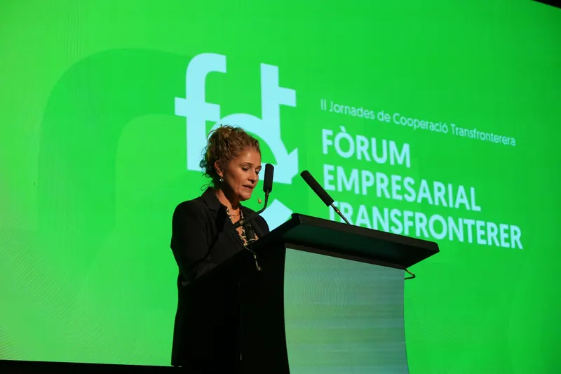 Una ponente habla en el atril ante la pantalla verde con el logotipo del Foro Empresarial Transfronterizo.